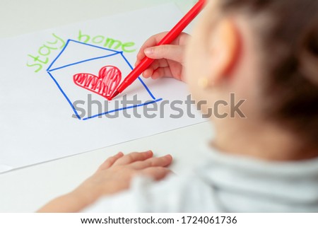 Stay Home concept. Child is drawing house with red heart on the sheet of paper with a written phrase over the house Stay Home on the easel.