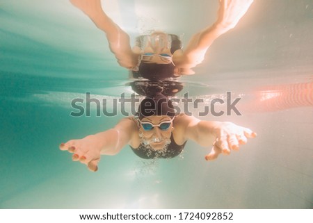 Female swimming instructor in a black swim suit, a cap and goggles is swimming under water