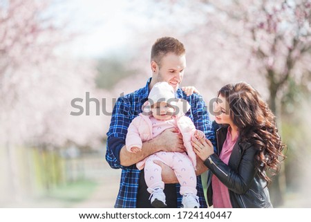 Young parents are holding in their arms a small pretty daughter. Beautiful family in a flowered garden. Sakura. Spring.