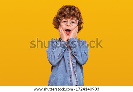 Surprised boy in denim jacket and glasses keeping hands near cheeks and looking at camera with opened mouth against yellow background