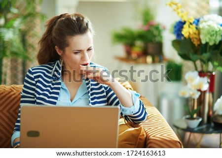 stressed trendy 40 years old housewife in blue blouse and striped jacket sitting on couch with laptop at modern home in sunny day.