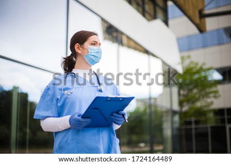 Young female caucasian UK doctor looking in distance with fear worry anxiety  uncertainty in eyes,wearing uniform  face mask,hope  faith  overcoming global Coronavirus COVID-19 pandemic crisis Royalty-Free Stock Photo #1724164489