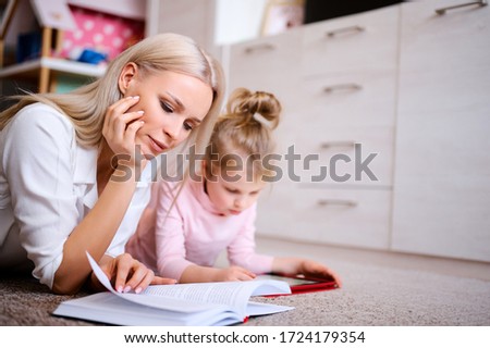 Young mother reads book at home lying on the floor with her daughter, child watching cartoons on digital tablet and playing on games, happy family, love, family concept.