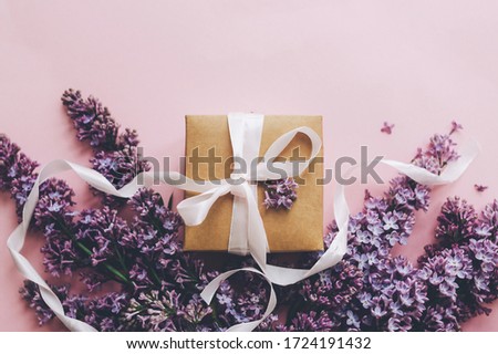 Lilac flowers and gift box on pink paper, stylish flat lay with copy space. Floral greeting card. Happy mothers day concept. Purple lilac flowers bouquet with craft present box with ribbon
