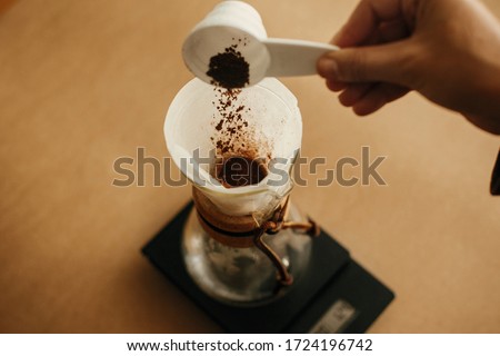 Hands pouring grounded coffee in filter. Preparing for alternative coffee brewing v60. Person holding spoon with grind coffee on background of glass kettle with pour over on scale Royalty-Free Stock Photo #1724196742