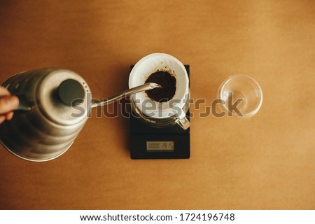 Pouring hot water from steel kettle in filter with ground coffee in pour over on scale, top view. Alternative coffee brewing v60. Barista making filter coffee on brown background Royalty-Free Stock Photo #1724196748
