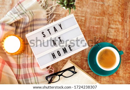 Board with Stay At Home writing, cup of coffee, burning candle and blanket on the wooden background. Lightbox with caution words. Corona virus, covid-19. Care and save concept