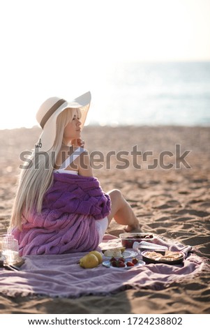 Young blonde woman having picnic at sandy beach over sea background in sun light. Wearing straw hat, dress and knitted sweater. Looking away. Summer season. Breakfast time. 