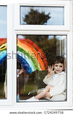 Little boy painting rainbow on a window and looking through the window while sitting home during quarantine. Let's all be well.