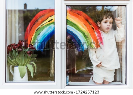 Little boy painting rainbow on a window and looking through the window while sitting home during quarantine. Dirty hands. Let's all be well.