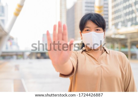 Overweight Asian woman with mask showing stop gesture in the city