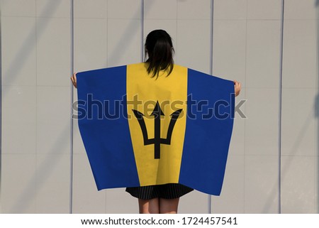 Woman is holding Barbados flag on her shoulder and turn back on grey background.