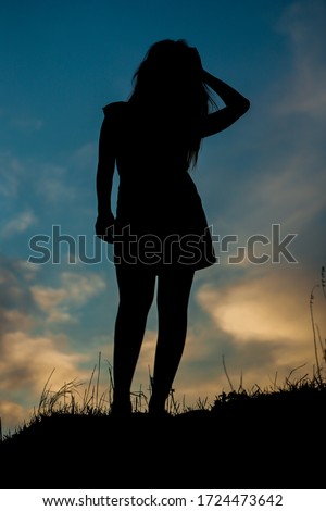 Silhouette of a tall girl against the sky with an evening sunset. A girl in a skirt stands on a mound with long hair and a short skirt.