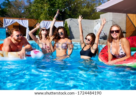 Group of friends have pool party dancing with inflatable floaty, watermelon swim ring toys. Attractive hot pretty women in bikini and sunglasses have fun relaxing and chilling on sunny summer day.
