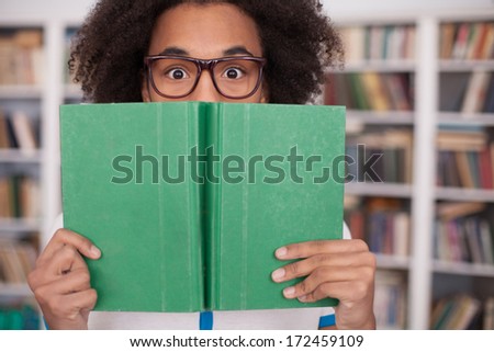 Worried about his exams. Terrified African teenager looking out of the book while standing in front of the book shelf