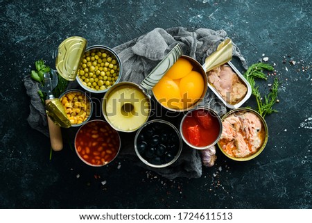 Canned vegetables, beans, fish and fruits in tin cans on black stone background. Food stocks.