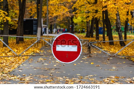 Stop sign at the gate for entering the park with autumn trees and fallen bright yellow leaves on the background of blurry people in the distance 