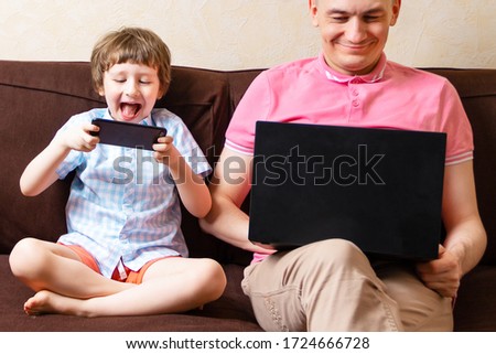 Happy young father sit on couch using laptop relax with kid son holding smartphone have fun together, smiling dad and little boy child enjoy weekend at home rest sofa busy with gadgets and play games.