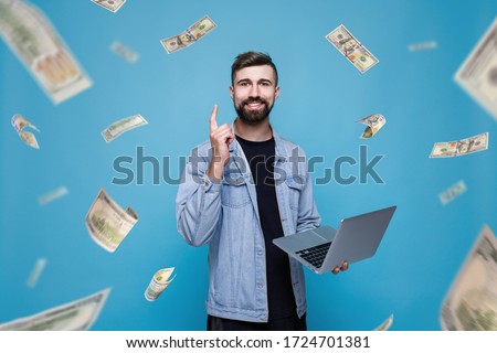 Young cheerful man with laptop in his hand. Guy browsing internet on smartphone and expresses happiness. Blue background. Money rain. Falling hundred dollar banknotes.
