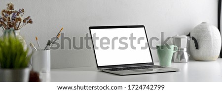 Side view of modern office desk with blank screen laptop, painting tools and decorations on white table