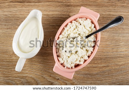 White sauceboat with sour cream, spoon in pink glass bowl with defatted grained cottage cheese on wooden table. Top view Royalty-Free Stock Photo #1724747590