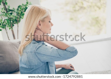 Closeup profile photo of unwell displeased blond mature lady sit couch stay home quarantine hold hand on neck suffer cramp spasm touch massaging injured pain place living room indoors