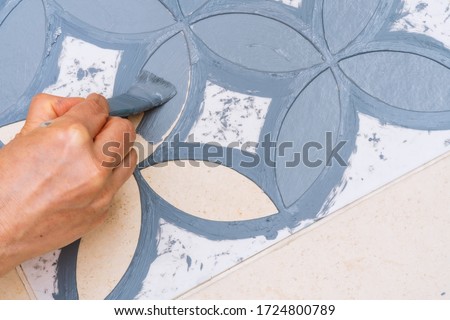 Stay at home and home improvement concept: Close up and top view of a hand holding a brush is painting a decorative template on the floor tiles into gray by using a vintage pattern stencil Royalty-Free Stock Photo #1724800789