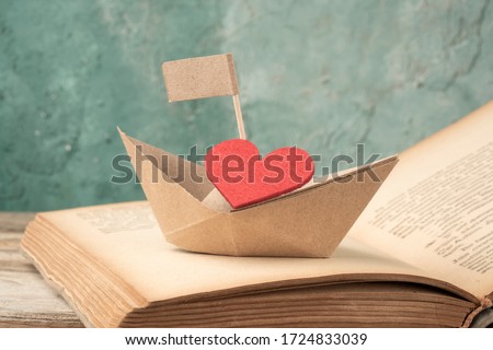 Folded paper sailing boat and red heart on old opened book on table. Hand crafted origami paper sailing boat on opened book pages, decorated with red hearts. valentines day or holiday concept photo