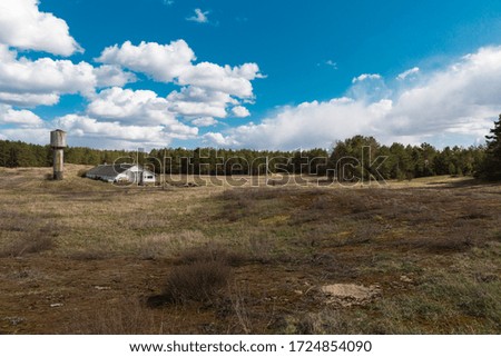 White farm, Old water tower, green forest, early spring. Clouds over the farm. Big meadow. Pine trees, earth, blue sky.
