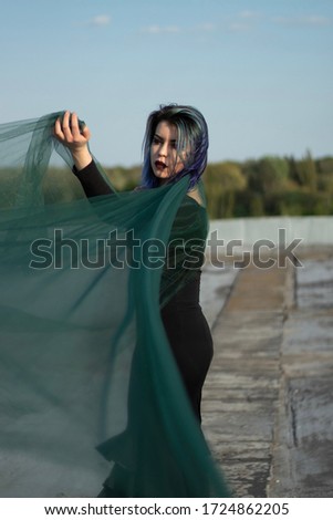 girl in black with blue hair with beautiful make-up swirls and dances with a blue transparent fabric on the roof against a blue sky with clouds