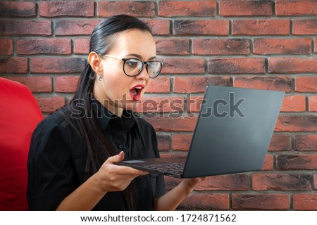 Beautiful young businesswoman in glasses and a black shirt looks at her laptop in surprise and says on her smartphone