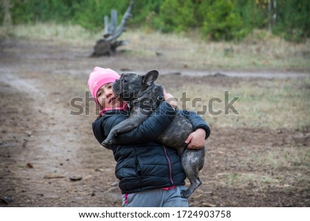 In the autumn forest, a little girl holds a gray French bulldog dog in her arms.