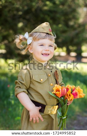 Portrait of a cute little girl in uniform with flowers on a green background. Victory Day, May 9 holiday.
