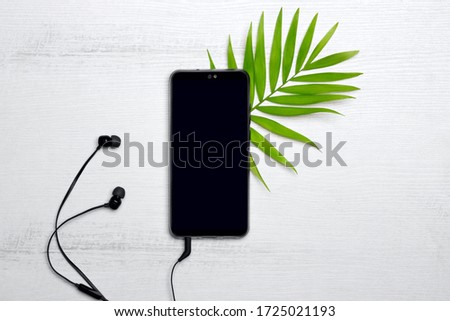 Mobile phone with headphones with green tropical palm leaf on white wooden background. Free copy space for text. Top view. Flat lay.