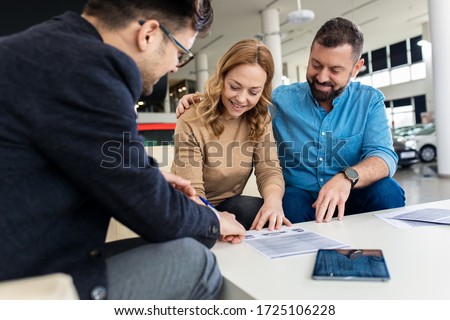 Mid-adult couple talking with sales agent and signing contract for new car in modern car showroom.