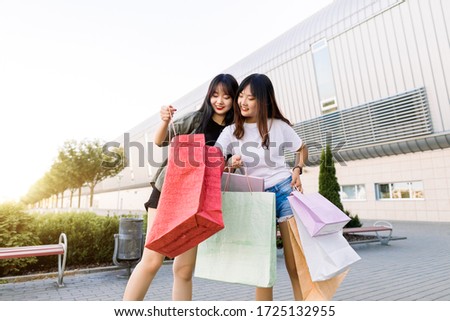Two happy smiling charming 25s asian women showing each other their shopping bags inside, standing on the shopping store building background outdoors in the city. Travel and shopping concept