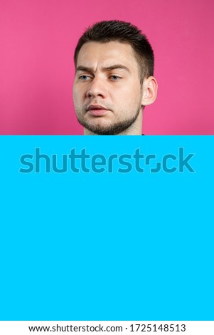 Young handsome man holding a blank cardboard blue banner over an isolated pink background, serious face, thinking about a question. Place for an inscription. Idea for recording thoughts