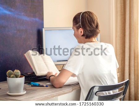 Teenage girl makes homework on online lesson learning at school. Back view of school girl reading book for distance education. Home schooling, social distancing, selective focus Royalty-Free Stock Photo #1725160330