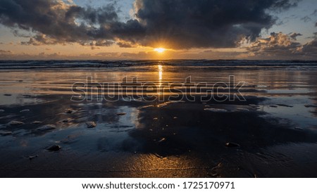 Beautiful sunset peaks through the clouds and reflects off the sand, as the waves gently roll into the foreground. 
