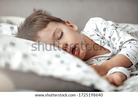 Little blond boy sleeping in his bed. Restless sleep of a baby with a cold. The kid sleeps with his mouth open. My little boy sleeping by the light of the window.