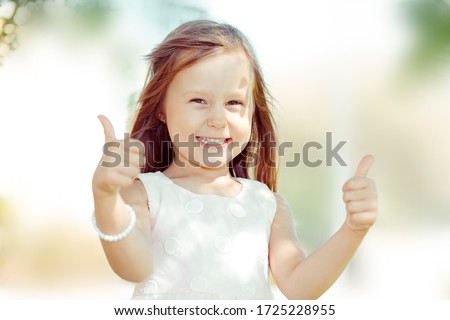 The little girl happy toddler kid showing thumbs up gesture with hands,  outdoors green tree park on background. Funny picture