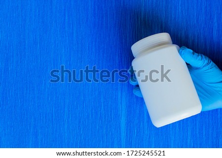 Hand in blue sterile gloves holds a white plastic jar for pills, medical treatment concept for diseases, blue background. Gloved hand with a medicine