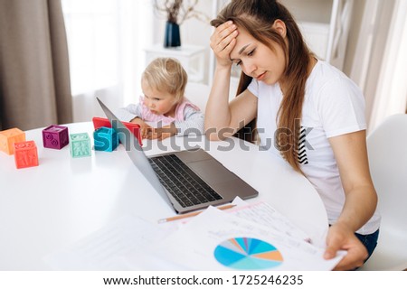 Work at home. Tired mom works remotely on laptop at home, and her little adorable daughter is sitting nearby and watching her favorite cartoon on a telephone