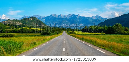A long straight road leading towards a mountains in France. Amazing bright colorful spring and summer landscape. Yellow fields of flowering rape and blue sky with clouds. Natural landscape, Europe. Royalty-Free Stock Photo #1725313039