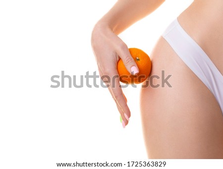 Female hip with smooth perfect skin in panties, close up. Caucasian woman holds an orange in hand and compare a peel with her body, isolated on white. Cellulite, skincare, spa, daily treatment