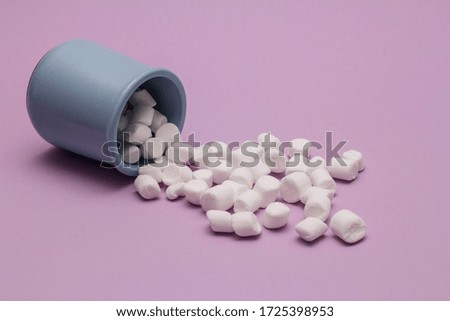 white marshmallows spilled from a blue earthenware Cup lying on its side on a pink paper background