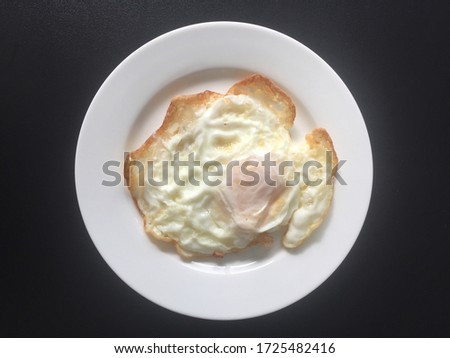 A white fried egg with a yolk in the middle The outer edge is brown, showing the crispiness of fried eggs. Place on a small white tile plate. Take a picture from above. Black background. top view.