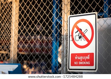 "No smoking" plate sign with Thai word which is located on the Chemical storage room's fence. Industrial safety sign photo, close-up and selective focus.