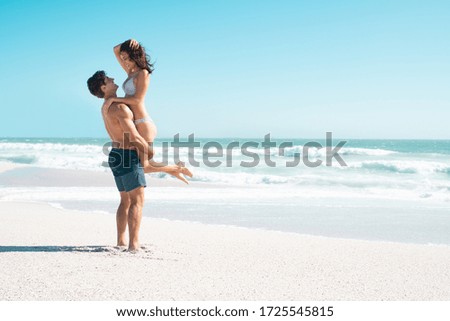 Loving couple hugging on the beach with copy space. Happy boyfriend in swinwear lifting his smiling girlfriend with ocean in background. Handsome husband carrying his beautiful wife in bikini.