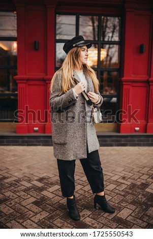a stylish girl on a background a beautiful red facade is dressed in a grey overcoat jeans leather shoe on a heel and with a head-dress by a hat by a cap and white bag, street style fashion